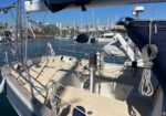 View from the deck of a sailboat at a marina: the helm wheel, coiled lines, and rigging in the foreground, with moored boats and city buildings in the background.