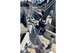 Close-up of a sailboat's bow rigging: winches, lines, and cleats with moored boats in a marina.