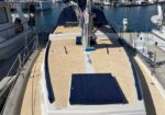Stern deck of a sailboat in a marina, tan deck with a dark blue hatch cover and blue trim along the edges, water and other boats in the background