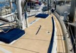 Sailboat deck with tan non-slip surface, blue cushions, and rigging, docked at a marina.