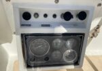 Boat cockpit instrument panel with several round gauges (tachometer and engine gauges) and labeled switches on a light gray console; sun glare on the glass.