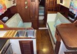 Wooden boat cabin with a galley on the left (double sink and stove), cushioned benches along both sides, and a narrow corridor leading to the forward cabin.