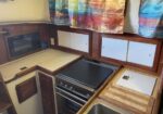 Compact boat galley with a stove/oven, microwave, wooden cabinets, beige countertops, a sink with a cutting board, and colorful curtains.
