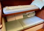 A compact boat cabin with an upper bunk having a blue pillow and light blanket, and a lower bunk with green cushions and folded linens under the shelf.