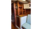 Wooden boat galley area with open shelves, a circular porthole window, and a white fridge or icebox beside a light blue cushioned seat.