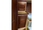 Wooden cabinet with louvered doors in a narrow boat cabin, light shelves above and a cushioned seating area nearby