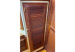 Tall wooden louvered cabinet door with brass hinge in a narrow interior space, likely closet storage.