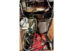 Red compact engine with hoses and wiring inside a wooden enclosure; battery and other components visible nearby.