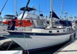 Sailboat docked at a marina with a white hull and blue stripe, an orange kayak on the bow, and other boats in the background.