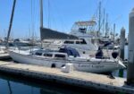 White sailboat named 'Latest Caper' docked at a marina on a sunny day, with masts and other boats in the background.