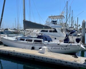 White sailboat named 'Latest Caper' docked at a marina on a sunny day, with masts and other boats in the background.