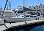 White motor yacht docked at a marina, with masts and neighboring boats, calm blue water, and a building backdrop under a clear sky.