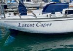 Sailing yacht named 'Latest Caper' moored in a marina with its hull visible above turquoise water.