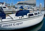 Sailboat with white hull and blue cabin, name 'Latest Caper' on the stern, docked in a marina with other boats.