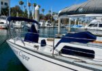 White sailboat docked in a marina with blue canvas covers, name partially visible on the hull, surrounded by other moored boats and palm trees under a clear blue sky.]