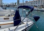 Sailboat moored in a marina with blue canvas covers on the boom and cockpit, calm water and buildings in the background.