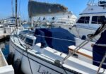 A white sailboat docked at a marina with blue canvas covers and other boats nearby.