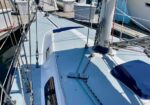 Sailboat deck at a marina with rigging, lines, rails, and a blue hatch cover; sunlight on the light-blue surface.