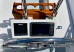 Boat helm with two navigation displays (B&G left, Garmin right), a polished stainless steering wheel in the foreground, and a wooden cup holder behind on a sunny deck.