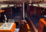 Interior of a sailboat cabin with a wooden galley, sink, and seating area on both sides, sunlight through windows.