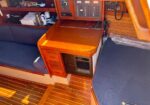 Wooden boat cabin featuring a small nav station with a foldable chart desk, blue cushioned benches, and a compact fridge beneath the desk under warm lighting.