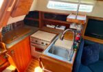 Boat galley with stainless sink, small oven/stove, and wooden cabinets; window above and blue cushioned seating nearby.