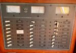 Marine electrical control panel with many labeled toggle switches and two gauges at top for DC distribution and battery status