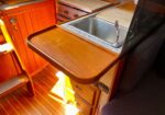 Wooden fold-out counter beside a stainless steel sink in a boat galley/kitchen area, with warm wood cabinetry around it