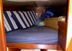 Boat cabin bunk with blue sheets, striped pillows, and cardboard boxes on the bed.