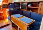 Inside a boat cabin: wood-paneled salon with blue cushioned seating, a table set with two blue placemats, and a small TV along the wall.