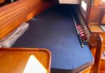 Wooden boat cabin with a blue cushioned berth and stored gear along the right, including a rolled striped item.