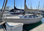White sailboat docked at a marina beside a white utility box, with masts and other boats in the background under a blue sky.