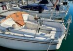 White sailboat docked in a marina with a wooden hatch cover, steering wheel, and metal railing in the cockpit, neighboring boats in the background