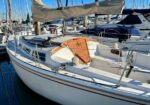 White sailboat docked in a marina with a tan sail cover, wooden companionway hatch, and stainless railings amid other moored boats.