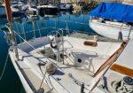 White sailboat cockpit with steering wheel, rigging, and coiled lines, docked in calm marina with other boats nearby