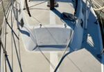 View down the center of a sailboat deck at a marina, with rigging, rope, and hatch in the foreground and calm water in the background.