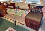 Interior of a boat cabin with a long beige bench and a tropical floral back cushion, a wooden cabinet with drawers, and a mounted radio/controls panel; a red fire extinguisher is on the wall and a small table sits beside the seating area.