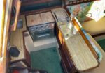 Compact boat galley with a small stove/oven, cutting boards, and a sink along a wooden counter; teal carpet and floral seating to the right.