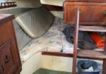 Interior of a small boat cabin with floral cushions and pillows, wooden cabinets and a ladder, cluttered with bags and gear.