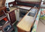 Boat galley with a narrow wooden counter, stainless sink, and a two-burner stove; cabinets and a cushioned seating area with coral-pattern upholstery on the right.