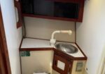 Compact boat head: white toilet, round sink with curved faucet, wood-toned cabinets, and a small overhead hatch/window above.