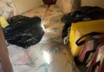 Cluttered boat cabin bed with floral cushions, black trash bags, and a yellow tote bag atop the mattress.