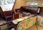 Interior of a boat galley with wooden cabinets, small sink, stove, and a colorful coral-pattern seating area; marina visible through windows.