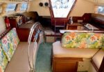 Interior of a sailboat cabin with cushioned seating and colorful coral-pattern upholstery, a circular steering wheel, and an open hatch to the deck ahead.