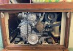 Small marine diesel engine mounted in a wooden cabinet with belts and hoses visible.