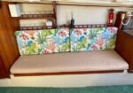 Inside a boat cabin with a beige bench seat and two colorful coral-pattern cushions, wooden shelving, a fire extinguisher, and a radio.