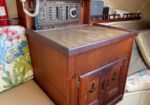 Wooden boat cabinet with a vintage radio on top, built-in storage doors, set between cushioned seating and a patterned chair in a small cabin.