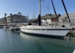 White sailboat moored at a marina, with other boats, calm rippled water, and waterfront buildings in the background.