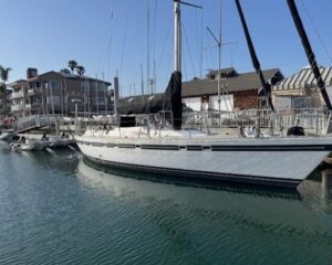 White sailboat moored at a marina, with other boats, calm rippled water, and waterfront buildings in the background.
