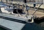 Close-up of a sailboat moored at a marina, showing the bow with an anchor and chain, deck hardware, and a wooden dock in the background.